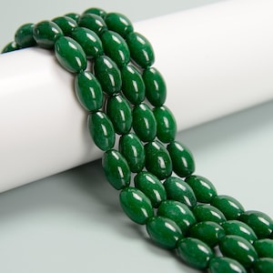 May include: Close-up of strands of polished, oval-shaped, deep green beads. The beads are strung together, creating a layered effect. The beads have a glossy finish, reflecting light. The background is a soft white and gray.