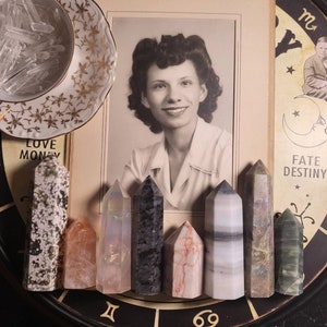 May include: A collection of seven crystal points, including a clear quartz point, a pink rose quartz point, a white and gray banded agate point, a black and gray obsidian point, a green aventurine point, a white and gray banded calcite point, and a brown and white jasper point. The points are arranged in a circle on a black surface with a zodiac symbol in the center.