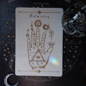 May include: A wooden plaque with the word "Palmistry" engraved on it. The plaque features a detailed illustration of a hand with various symbols and astrological signs, including a sun, moon, stars, and a crown.