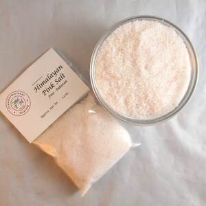 May include: A small clear plastic bag filled with fine pink Himalayan salt. The bag has a white label that reads "Himalayan Pink Salt Fine - Pakistan Approx. Net Wt. - 2.5 oz". The bag is sitting on a white surface next to a clear glass bowl filled with the same pink salt.