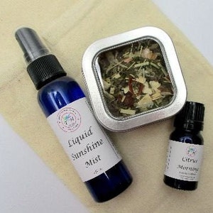 May include: A set of three aromatherapy products in a beige fabric pouch. The products include a blue glass bottle with a spray nozzle labeled "Liquid Sunshine Mist", a small tin container filled with dried herbs, and a dark glass bottle labeled "Citrus Morning Essential Oil".