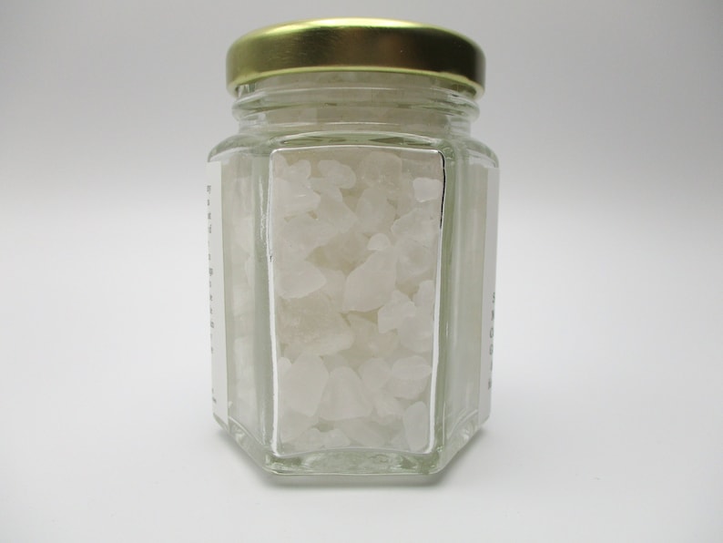 May include: A glass jar with a gold lid filled with white crystals. The jar is hexagonal in shape and has a label on the side.