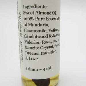 Op de afbeelding: Een klein, doorzichtig glazen flesje met een wit etiket. Het etiket vermeldt "Ingredi&euml;nten: Zoete amandel olie; 100% pure essenti&euml;le olie van mandarijn, kamille, vetiver, sandelhout en jasmijn, valeriaanwortel; en een kunzietkristal, zoete dromen intentie en liefde 1 dram ~ 4 ml".