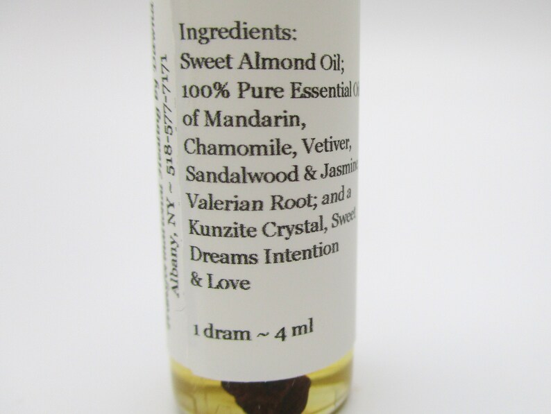 Op de afbeelding: Een klein, doorzichtig glazen flesje met een wit etiket. Het etiket vermeldt "Ingredi&euml;nten: Zoete amandel olie; 100% pure essenti&euml;le olie van mandarijn, kamille, vetiver, sandelhout en jasmijn, valeriaanwortel; en een kunzietkristal, zoete dromen intentie en liefde 1 dram ~ 4 ml".