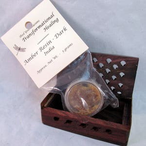 May include: A small, dark wooden box with carved details holds a clear plastic package containing amber resin. A white tag with the text "Transformational Healing" and "Amber Resin - Dark" is attached. The resin weighs approximately 5 grams.