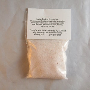 May include: A small clear plastic bag filled with white crystals. The bag has a white label with the text "Metaphysical Properties: Cleansing, purification, consecration, grounding, rituals, protection, great for cleansing crystals, love, marriage, reunion, new hope, healing, and bright journey Transformational Healing by Dawna Etsy.com/shop/DeesTransformHealing Albany, NY 518-577-7171".