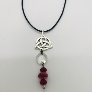 May include: A silver Celtic knot pendant necklace with a clear crystal and red crystal beads hanging from it. The necklace is on a black cord.