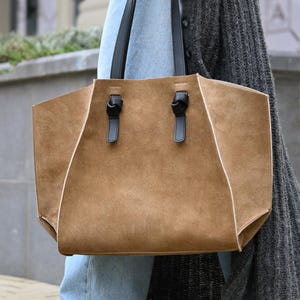 May include: A tan suede tote bag with black handles and accents. The bag has a geometric design with white stitching and is being carried by a person wearing blue jeans and a gray cardigan.