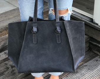 May include: A large, dark gray leather tote bag with knotted handles. The bag is held by a person wearing blue jeans.