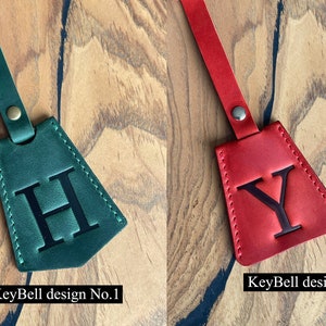 May include: Two leather keychains with a triangle shape and a single letter embossed on each. The keychain on the left is green with the letter "H" and the keychain on the right is red with the letter "Y". The text "KeyBell design No. 1" is below the green keychain and the text "KeyBell design No. 2" is below the red keychain.
