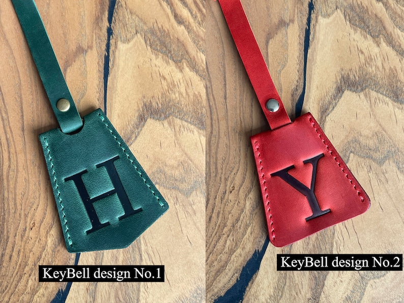 May include: Two leather keychains with a triangle shape and a single letter embossed on each. The keychain on the left is green with the letter "H" and the keychain on the right is red with the letter "Y". The text "KeyBell design No. 1" is below the green keychain and the text "KeyBell design No. 2" is below the red keychain.