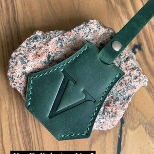 May include: A dark green leather key fob with a stitched border and a large, black, uppercase "V" in the center. The fob is attached to a brown leather strap with a silver metal rivet. The text "KeyBell design No. 5" is visible below the fob.
