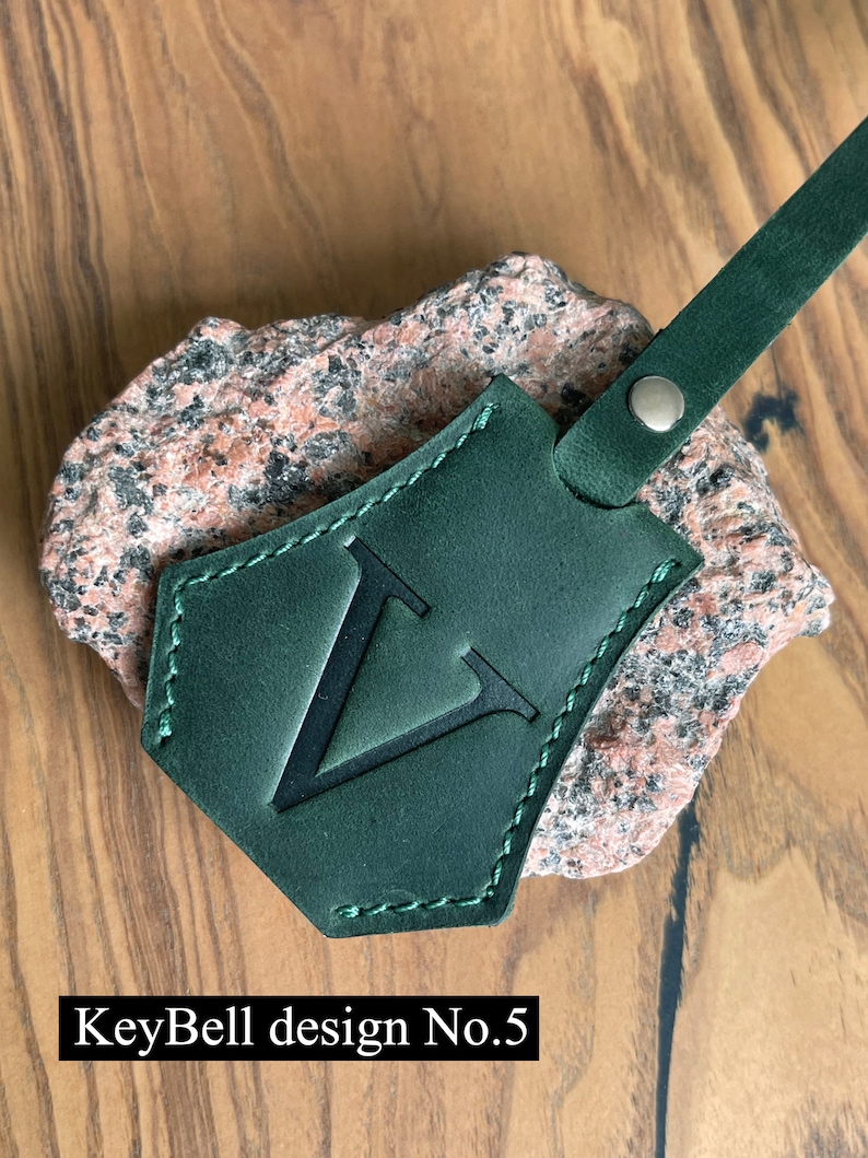 May include: A dark green leather key fob with a stitched border and a large, black, uppercase "V" in the center. The fob is attached to a brown leather strap with a silver metal rivet. The text "KeyBell design No. 5" is visible below the fob.