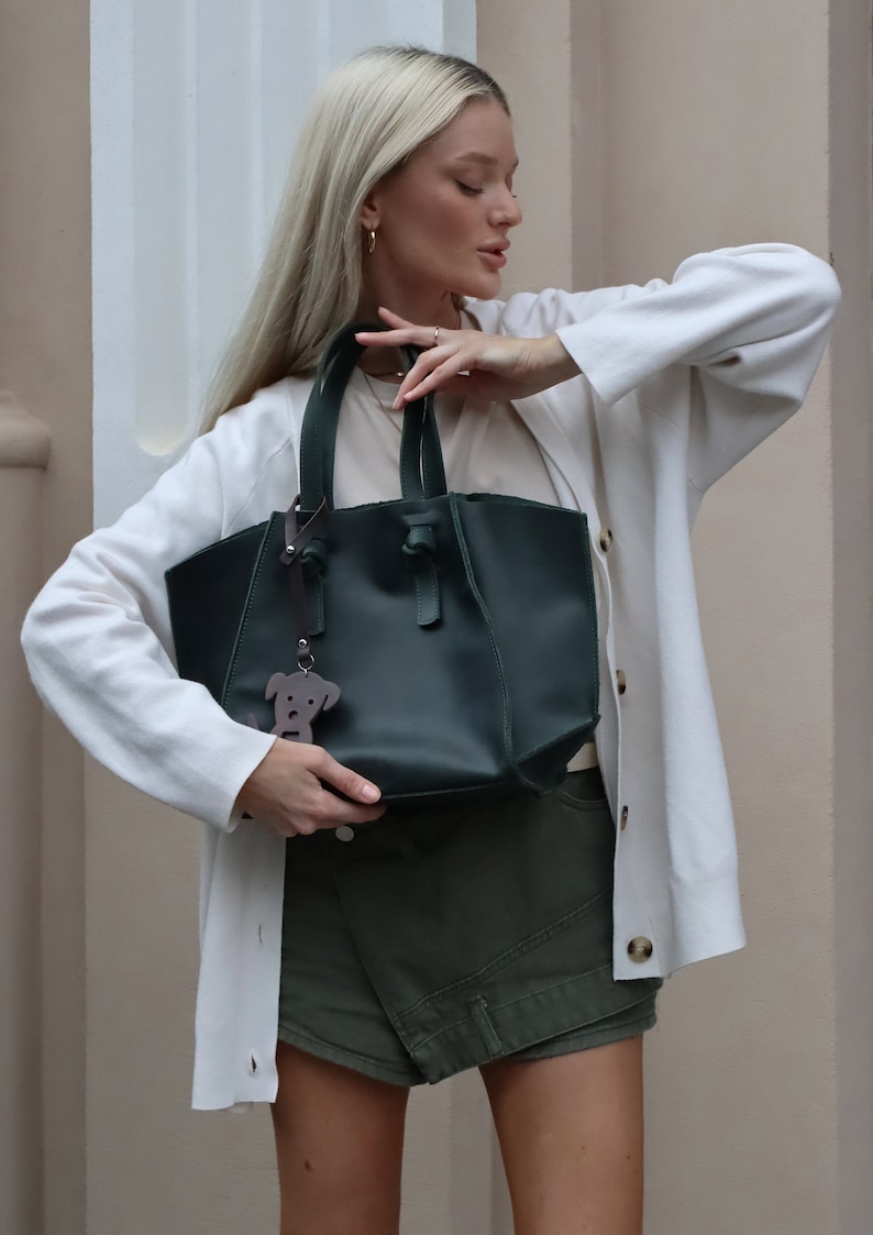 Puede incluir: Una mujer con un c&aacute;rdigan blanco y pantalones cortos verdes sostiene un bolso tote de cuero verde oscuro con un dije de perro de cuero marr&oacute;n.