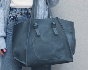 Puede incluir: Un bolso tote grande de cuero azul con un bolsillo frontal. El bolso tiene una parte superior ancha y abierta y dos asas.