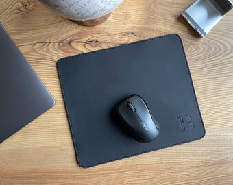 Könnte beinhalten: Schwarze Leder-Mausmatte mit einem genähten Rand und einem Monogramm mit dem Buchstaben "B" in der rechten unteren Ecke. Eine schwarze kabellose Maus liegt auf der Matte.