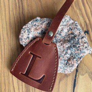 May include: Brown leather key chain with a stitched edge and a large letter L embossed on the front. The keychain is attached to a brown leather strap with a silver rivet. The text "KeyBell design No. 8" is printed below the keychain.