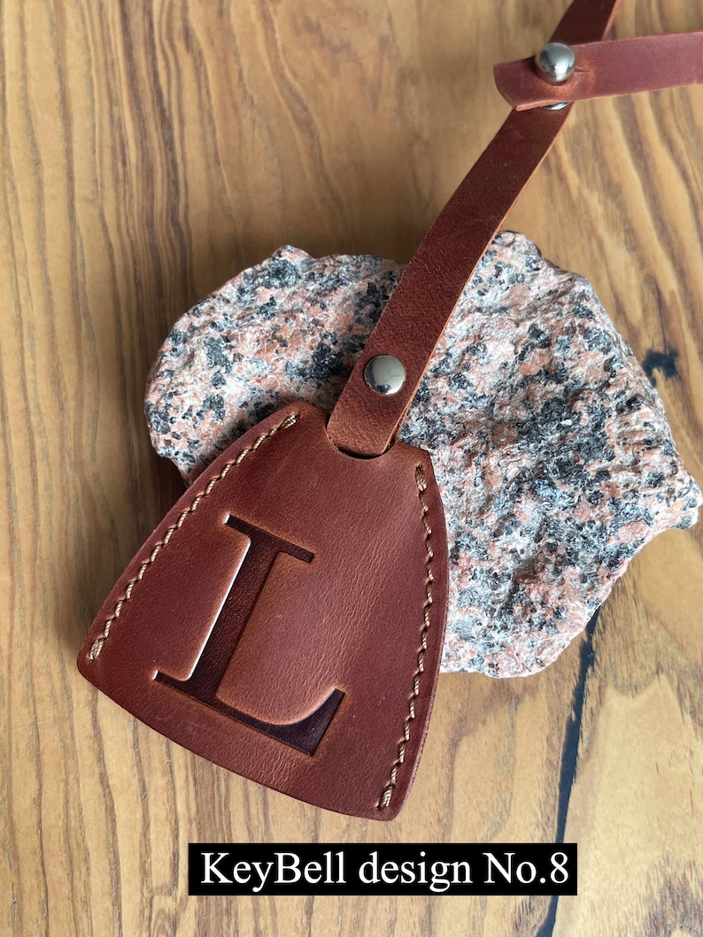 May include: Brown leather key chain with a stitched edge and a large letter L embossed on the front. The keychain is attached to a brown leather strap with a silver rivet. The text "KeyBell design No. 8" is printed below the keychain.