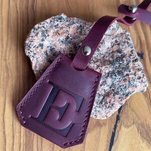 May include: Burgundy leather key chain with a stitched edge and a large letter "E" embossed on the front. The keychain has a metal loop and a metal snap closure. KeyBell design No. 6.