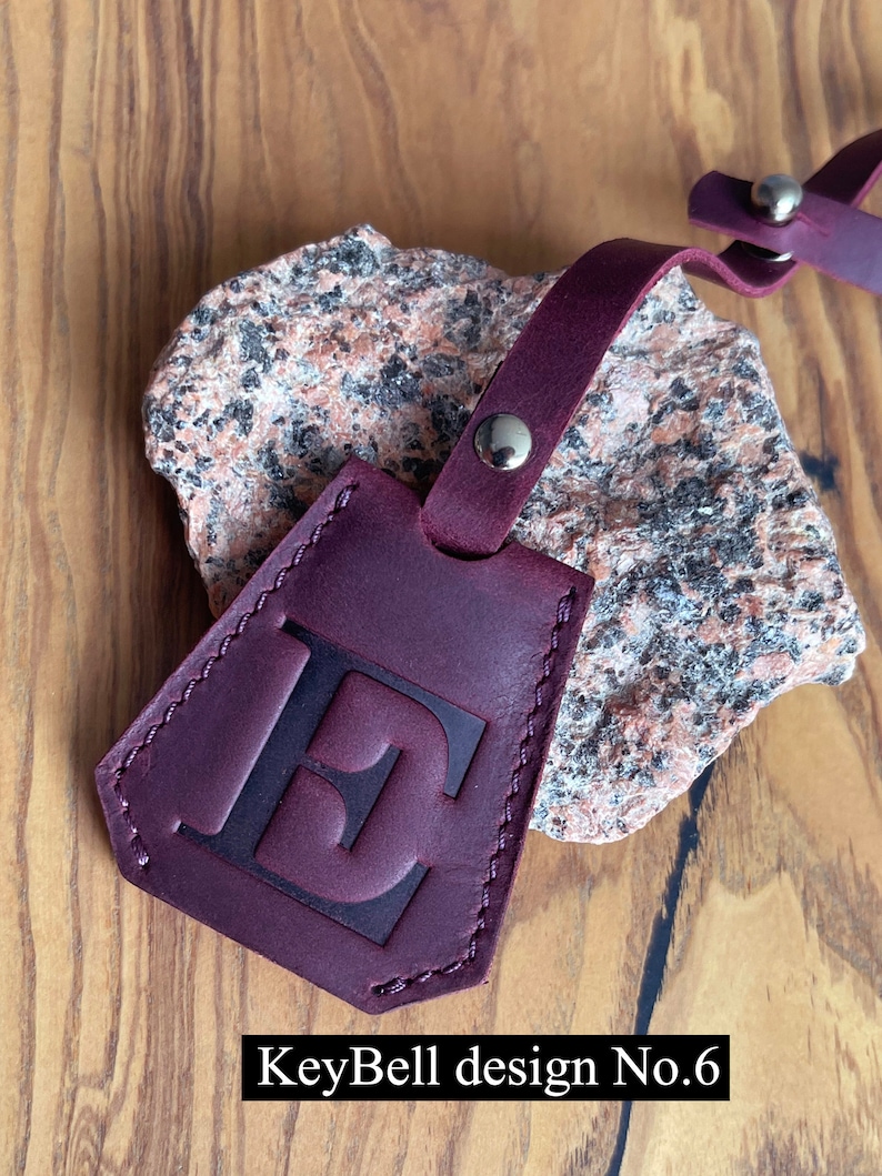 May include: Burgundy leather key chain with a stitched edge and a large letter "E" embossed on the front. The keychain has a metal loop and a metal snap closure. KeyBell design No. 6.