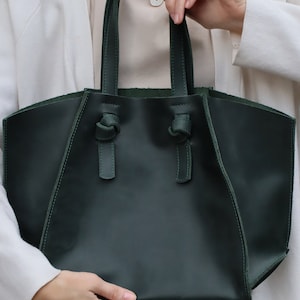 Puede incluir: Un bolso tote de cuero verde oscuro con asas anudadas. El bolso tiene un dise&ntilde;o simple y elegante.