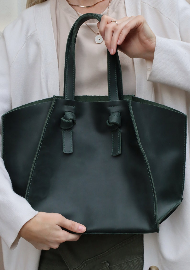 Puede incluir: Un bolso tote de cuero verde oscuro con asas anudadas. El bolso tiene un dise&ntilde;o simple y elegante.