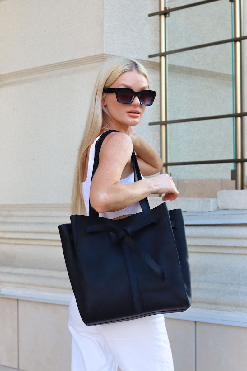 May include: A black leather tote bag with a knotted strap detail. The bag is being held by a person wearing a white tank top and sunglasses.