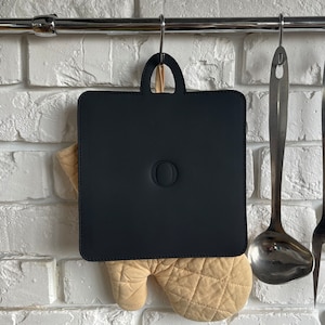 Op de afbeelding: Een zwarte ovenwant met een gestikt ontwerp hangt aan een rek met twee metalen keukengerei. De ovenwant heeft een ronde uitsparing in het midden.