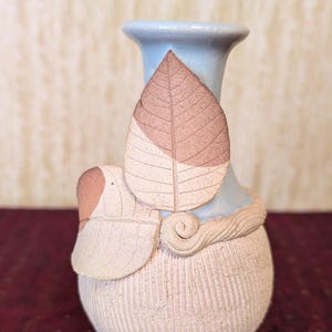 Op de afbeelding: Een keramische vaas met een lichtblauwe hals en een getextureerde, gebroken witte body. Twee gestileerde bladeren in bruin- en crèmetinten zijn aan de vaas bevestigd. De vaas heeft een unieke, handgemaakte uitstraling.