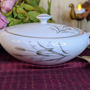 May include: White porcelain serving dish with lid, featuring a wheat design in gray and gold trim. The dish has two handles and a knob on the lid. The background includes purple flowers and a decorative candle holder.