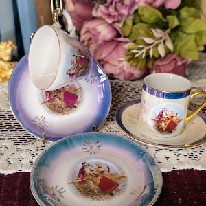 May include: A set of vintage porcelain teacups and saucers. The teacups and saucers have a blue and white iridescent finish with gold trim. Each piece features a painted scene of figures in period clothing. The set includes two saucers and two teacups.
