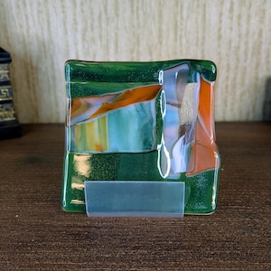May include: A square fused glass coaster featuring a deep green base with embedded shimmering particles. The surface displays abstract diagonal bands of orange, white, and translucent glass, creating a layered effect. It rests on a clear plastic stand.