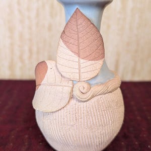 May include: A ceramic vase with a pale blue neck and a textured, off-white body. Two stylized leaves in shades of brown and beige are attached to the vase. The vase has a unique, handcrafted appearance.