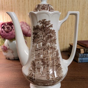 May include: A white porcelain coffee pot featuring a brown transferware print of a colonial-style building with a clock tower, surrounded by trees and figures in period clothing. The pot has a scalloped rim, a tall spout, and a matching lid.