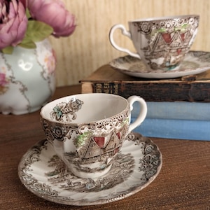 May include: Two ceramic teacups and matching saucers featuring a brown and white transferware pattern of a rural landscape with houses and figures. The cups have a scalloped rim and a curved handle, with subtle red and green hand-painted accents.