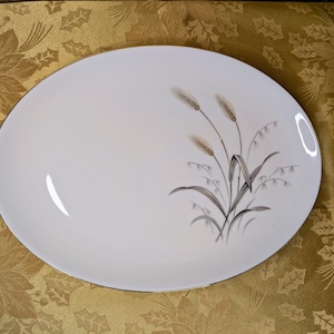 May include: An oval, white serving platter with a gold rim. The platter features a wheat stalk design in shades of brown, gray, and green. The platter is set on a gold floral tablecloth.