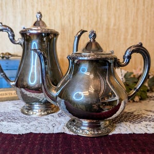 May include: Two antique silver tea pots with ornate handles and lids. The tea pots are highly reflective, with a polished finish. The tea pots are sitting on a white lace tablecloth.