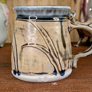 May include: A ceramic mug featuring a tan and cream glaze with dark blue drip accents. The mug has a sculpted handle with a decorative protrusion at the top and a smooth white base. The surface displays raised, flowing leaf-like patterns.