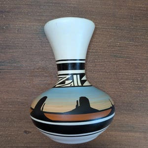 May include: A ceramic vase with a white neck and base, featuring a painted landscape of black rock formations against a blue and tan sky. The design includes black geometric patterns around the neck and a wide black band encircling the lower body.