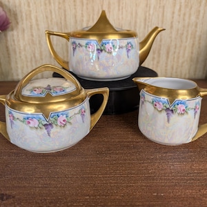 May include: A porcelain tea set comprising a teapot, a lidded sugar bowl, and a creamer. Each piece features an iridescent white finish, gold-toned handles and rims, and a decorative band of pink roses with green leaves and blue accents.