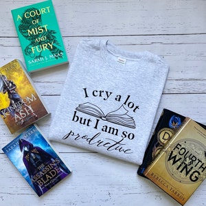 Puede incluir: Una vista plana de una camiseta gris con el texto "Lloro mucho pero soy tan productivo" y un libro abierto en la parte delantera. Hay cuatro libros de bolsillo sobre un fondo de madera blanca. Los libros son "A Court of Mist and Fury", "Kingdom of Ash", "The Assassin's Blade" y "Fourth Wing".