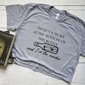 May include: A gray cropped t-shirt with a black graphic of a bottle with a rolled up message inside. The text on the shirt reads "There's a story at the bottom of this bottle and I'm the reader."