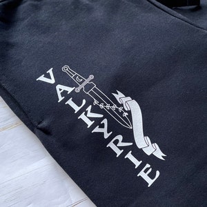May include: Black sweatpants with a white graphic design of a sword and the word "VALKYRIE" on the left leg.