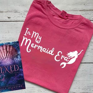 以下が含まれることがあります： 白い文字で「In My Mermaid Era」と書かれたピンクのTシャツ。Tシャツには白いシルエットのマーメイドも描かれている。Tシャツは白い木製の表面に置かれている。