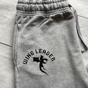 May include: Close-up of gray sweatpants with a drawstring waist. The text "WING LEADER" is printed in black, along with a graphic of a dragon. The sweatpants have a pocket on the side.