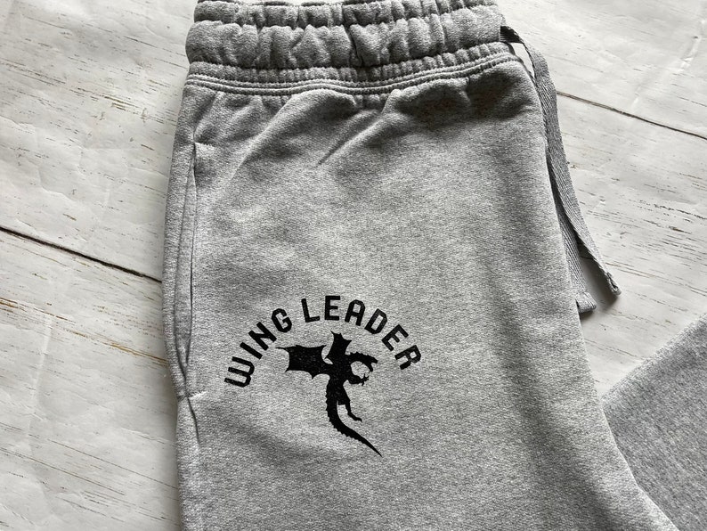 May include: Close-up of gray sweatpants with a drawstring waist. The text "WING LEADER" is printed in black, along with a graphic of a dragon. The sweatpants have a pocket on the side.