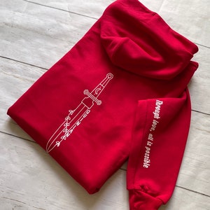 May include: Red hooded sweatshirt with a white sword design on the front and the text "Through love, all is possible" on the sleeve.