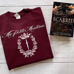 以下が含まれることがあります： 「Ma Petite Montueuse」と書いてある白いグラフィックデザインが施された、マルーンのトレーナー。王冠、剣、葉が描かれています。