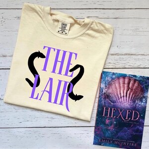 以下が含まれることがあります： クリーム色のTシャツに、紫色の文字で「THE LAIR」と書いてあり、黒いウナギのシルエットが2つ描かれています。Tシャツは白い木製の表面に置かれ、その隣には「HEXED」というタイトルのついた本が置かれています。本の表紙は紫色と青色で、貝殻が描かれています。