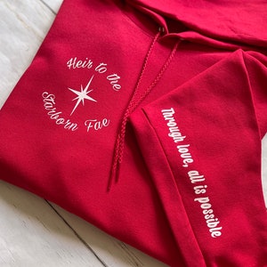May include: Red hooded sweatshirt with white embroidery. The front reads "Heir to the Starborn Fae" with a star design. The left sleeve reads "Through love, all is possible."